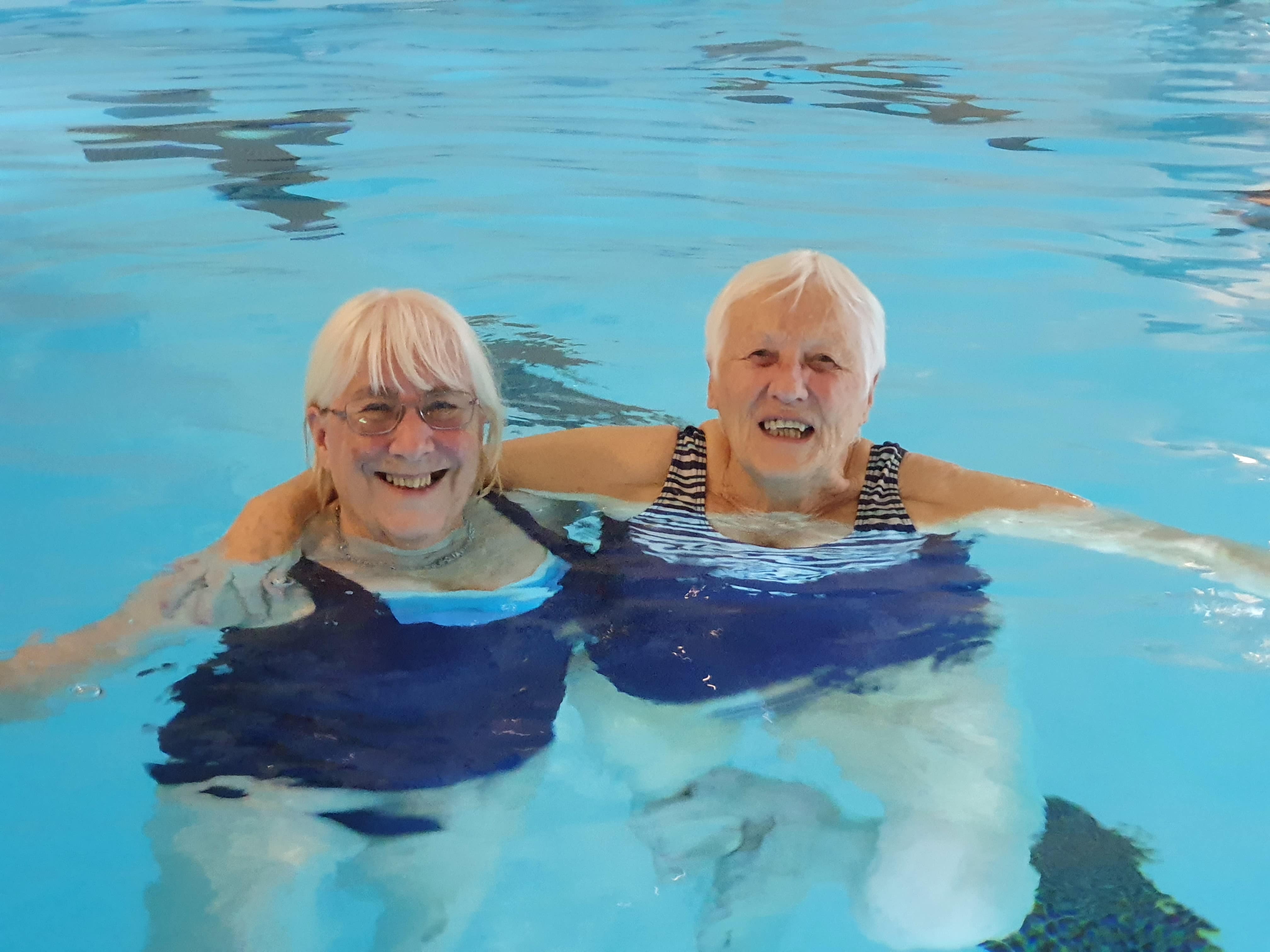 Two elderly women in a swimming pool with their arms around each other smiling 