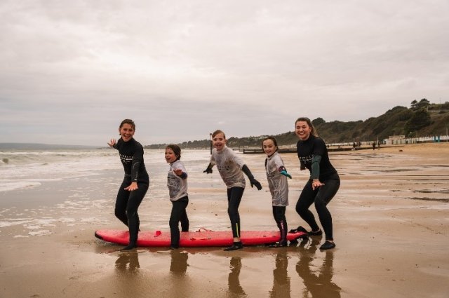 a group of 3 children and 2 adults in front of a surf board on the beach