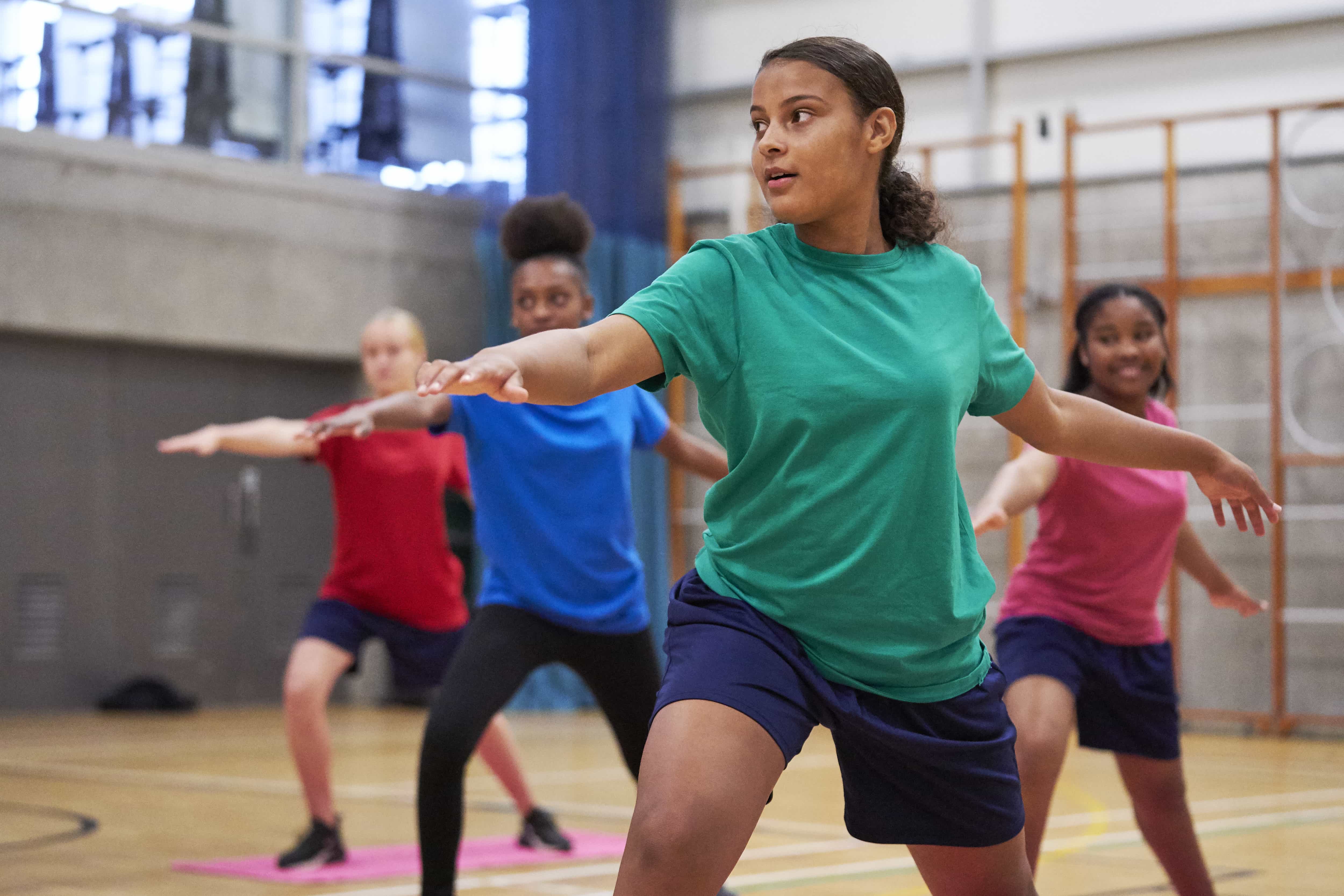 Young people wearing bright coloured t-shirts are taking part in a fitness class in a school sports hall 