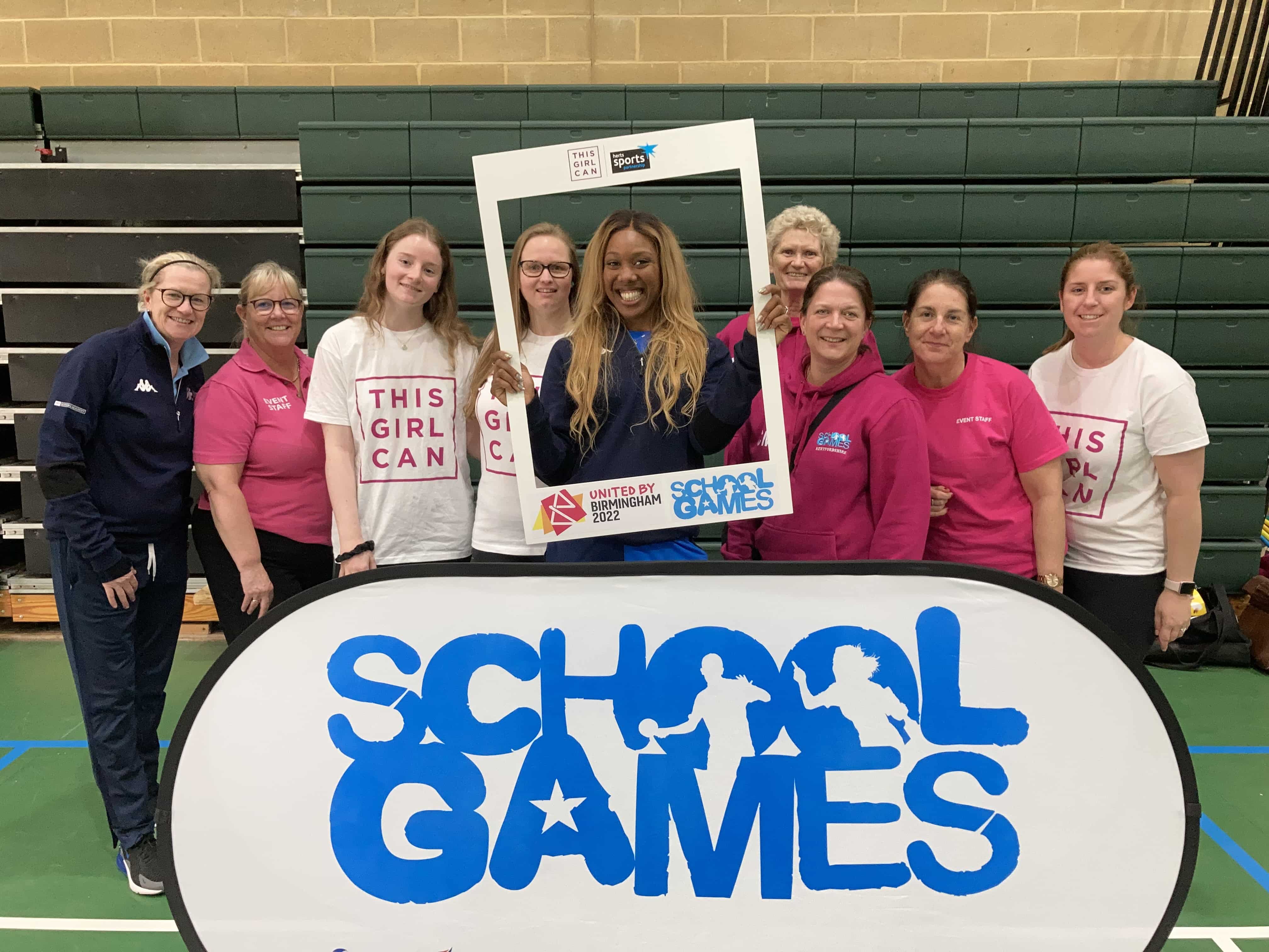 Group of girls with Montell Douglas standing behind a School, Games banner in a sports hall
