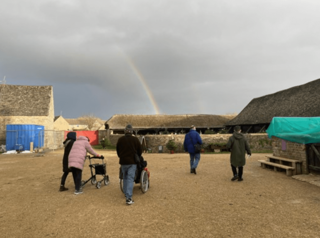 people walking across a yard with a rainbow in the background. One person is being pushed in a wheelchair