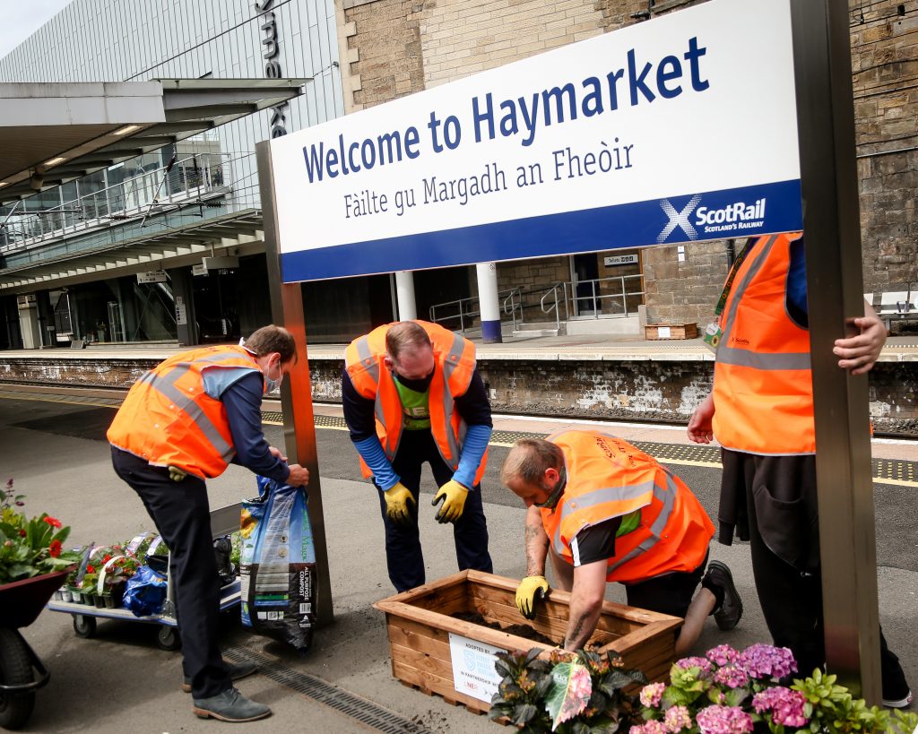 four men in high vis orange vests putting flowers in a rectangle planter on a railway station 