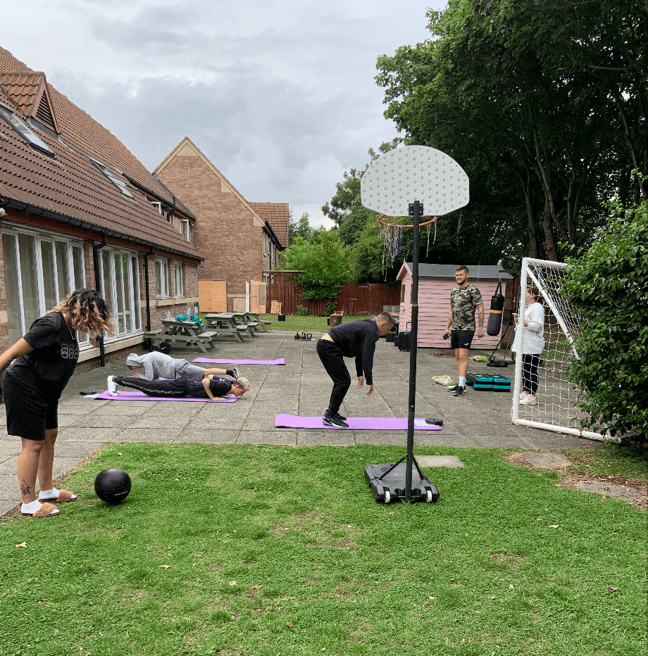 6 young adults taking part in a circuit session in a garden using medicine balls, yoga mats and basketball hoop