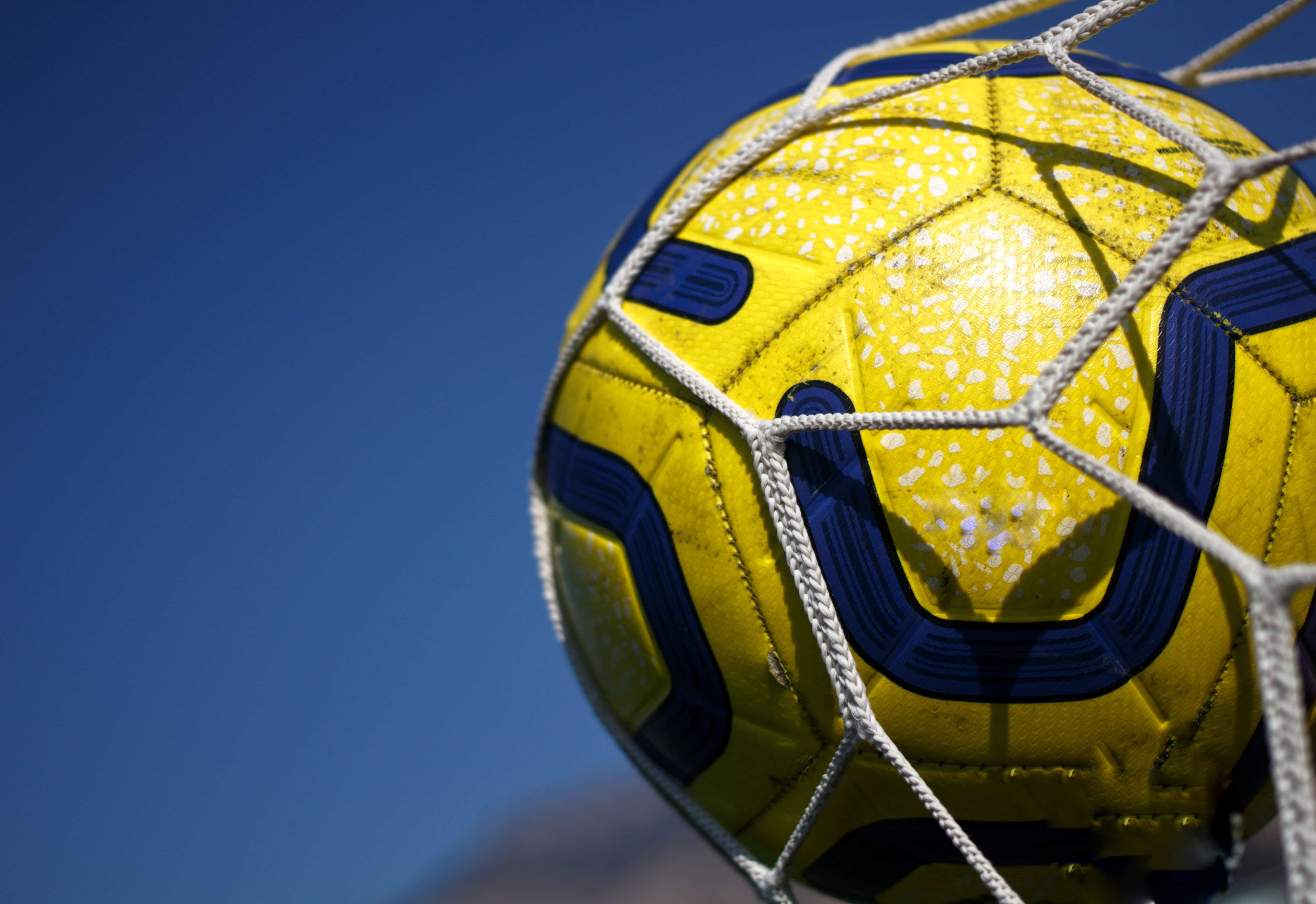 Yellow football with black swirl pattern in the net of a football goal against a blue sky. 