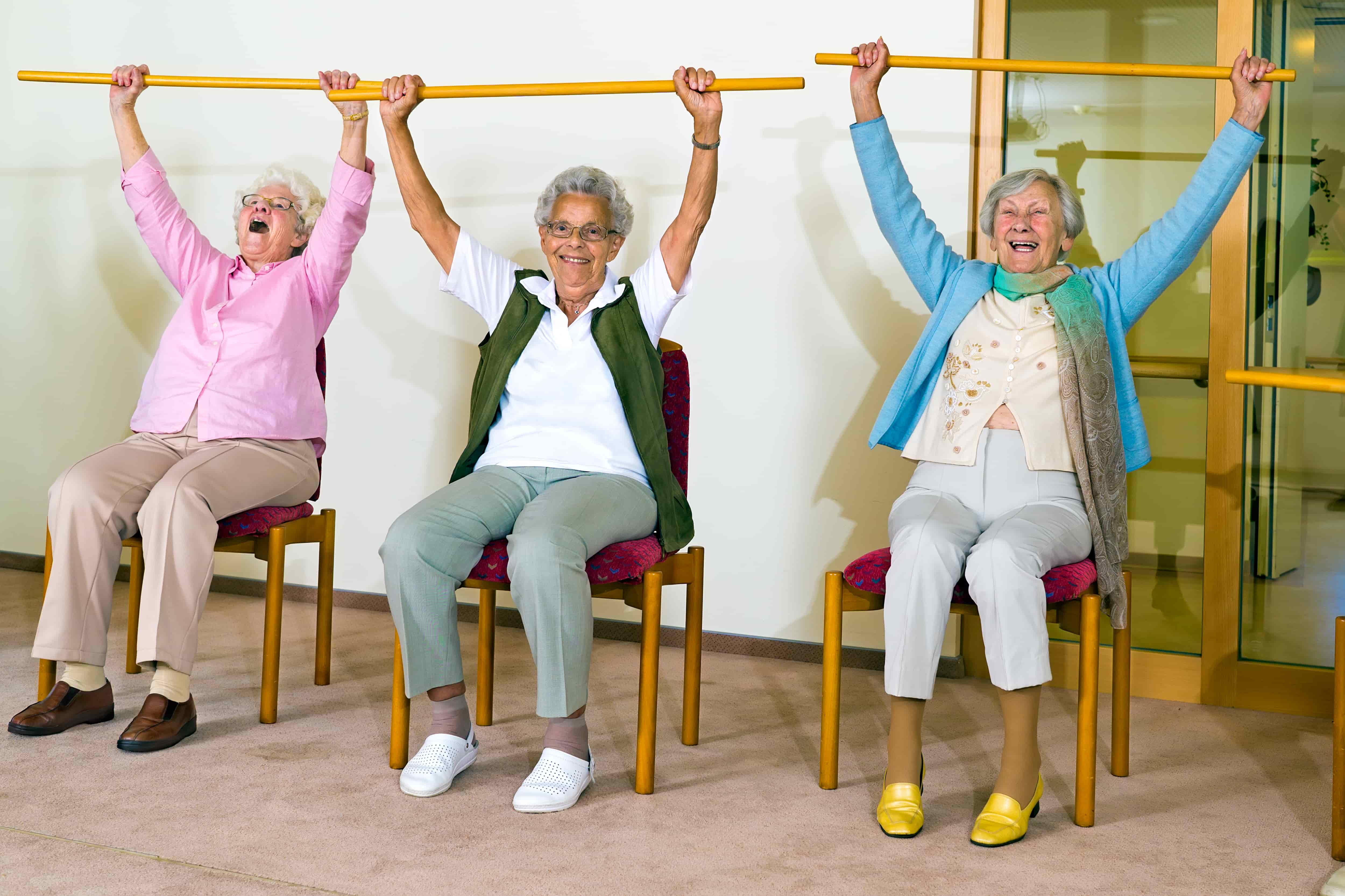 three older women smiling, sitting in chairs lifting wooden poles above their heads