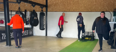 Image shows the inside of a boxing gym. There are punch bags hanging from the ceiling and three people are stretching to warm up. 