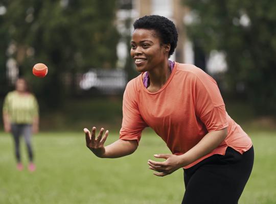 black female in active wear throwing a small orange ball in the park