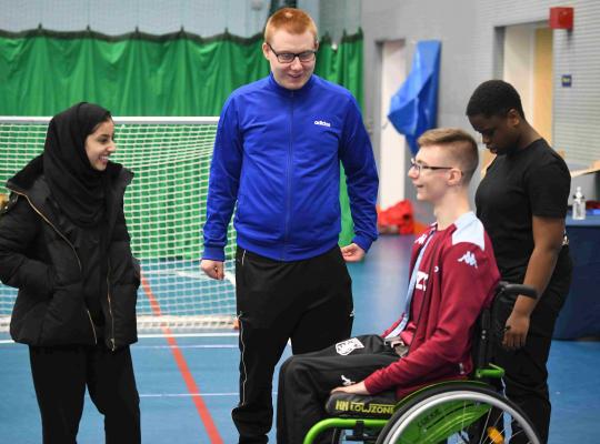 4 young leaders in a sports hall talking, one male is in a wheelchair and a female is wearing a hijab 