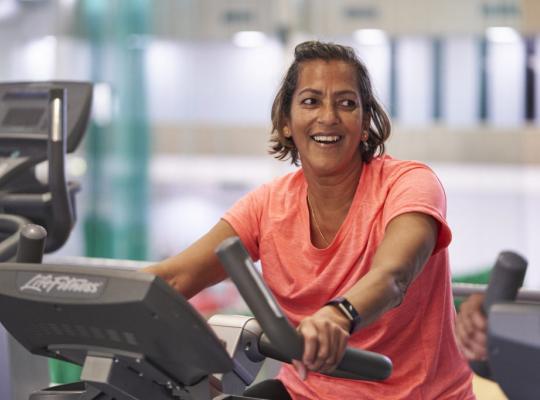 female in an orange t-shirt smiling whilst working out on a step machine in the gym