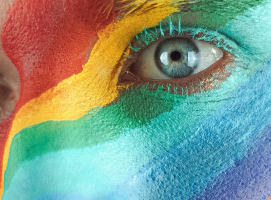 close up of a persons face with a rainbow colours painted over the bridge of the nose and right left eye