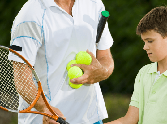 male coach showing boy how to hold a tennis racket whilst holding two tennis balls in other hand.