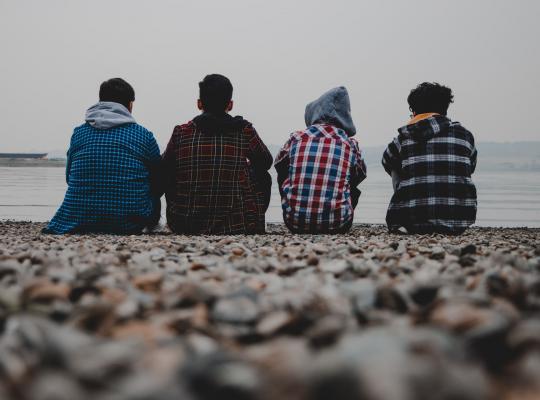 4 teenagers sitting on a pebble beach looking out to sea. 