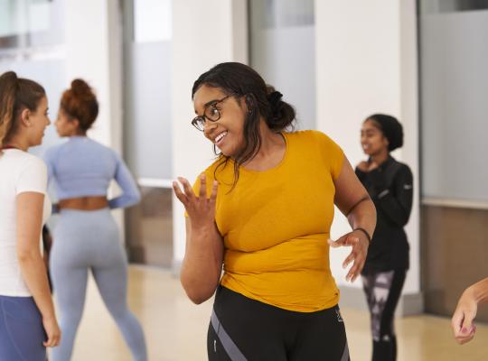 group female exercise class in an indoor space. Black female wearing a yellow top laughing at the front as she performs a dance move