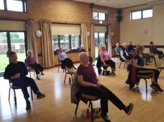 an older group of people doing chair based activity in hall