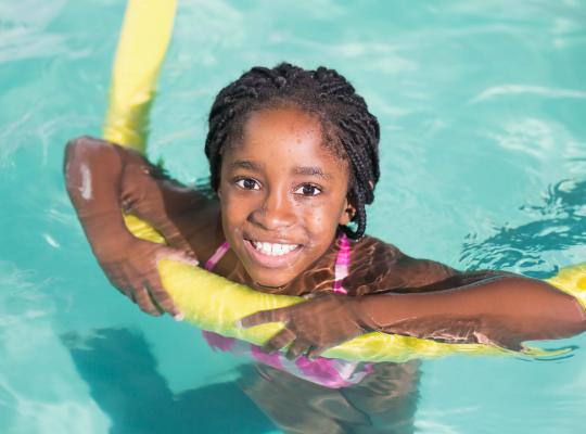 black girl smiling in a swimming pool holding a yellow long float 
