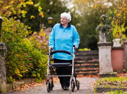 Older lady in blue fleece walking with a walking frame in a garden 